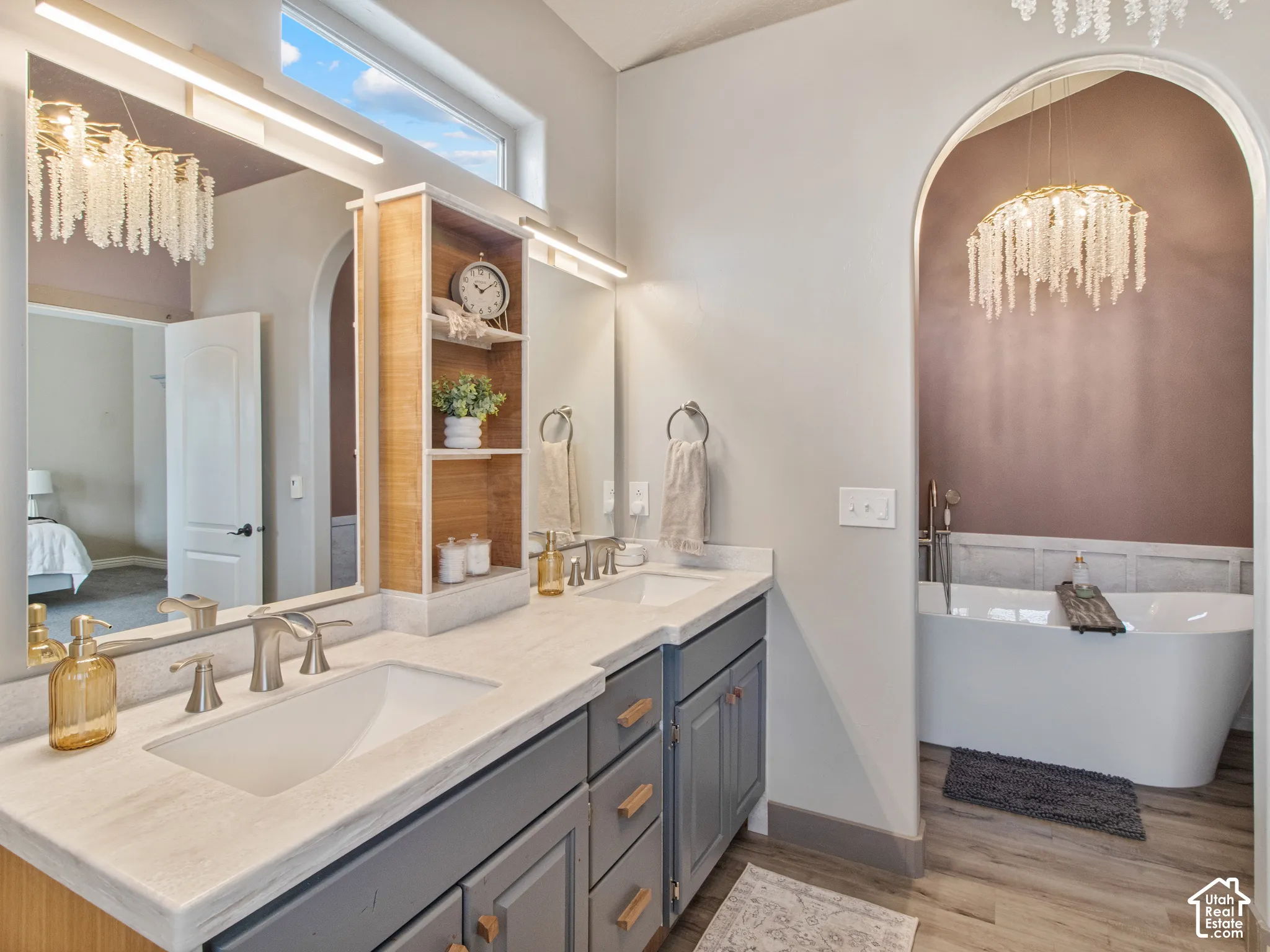Dual vanity with undermount sinks and modern faucets, integrated storage cabinetry, built-in shelving, freestanding tub, and wood-finish flooring
