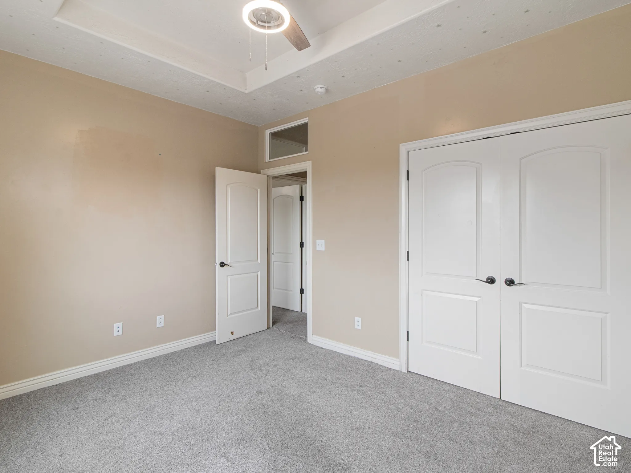 Tray ceiling with a ceiling fan and integrated lighting, neutral wall tones, gray carpeting, white bi-fold closet doors, and white trim