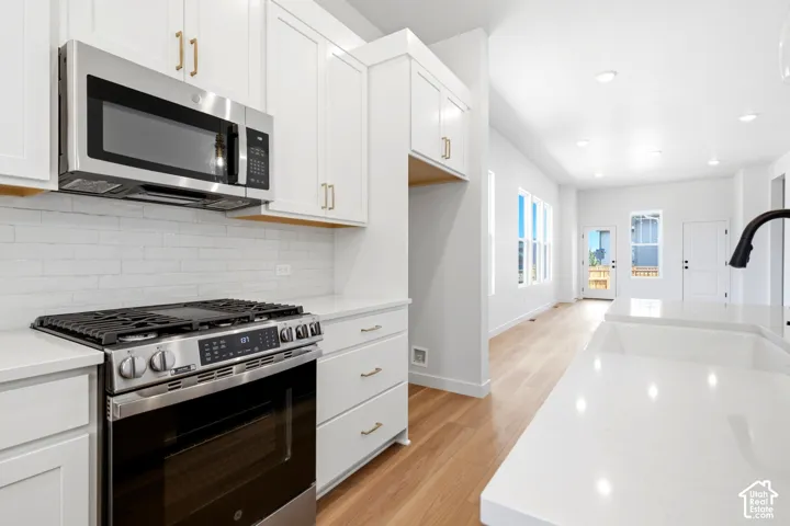White shaker cabinetry with gold-tone hardware, stainless steel gas range, white subway tile backsplash, white countertop island, and wood-finish flooring