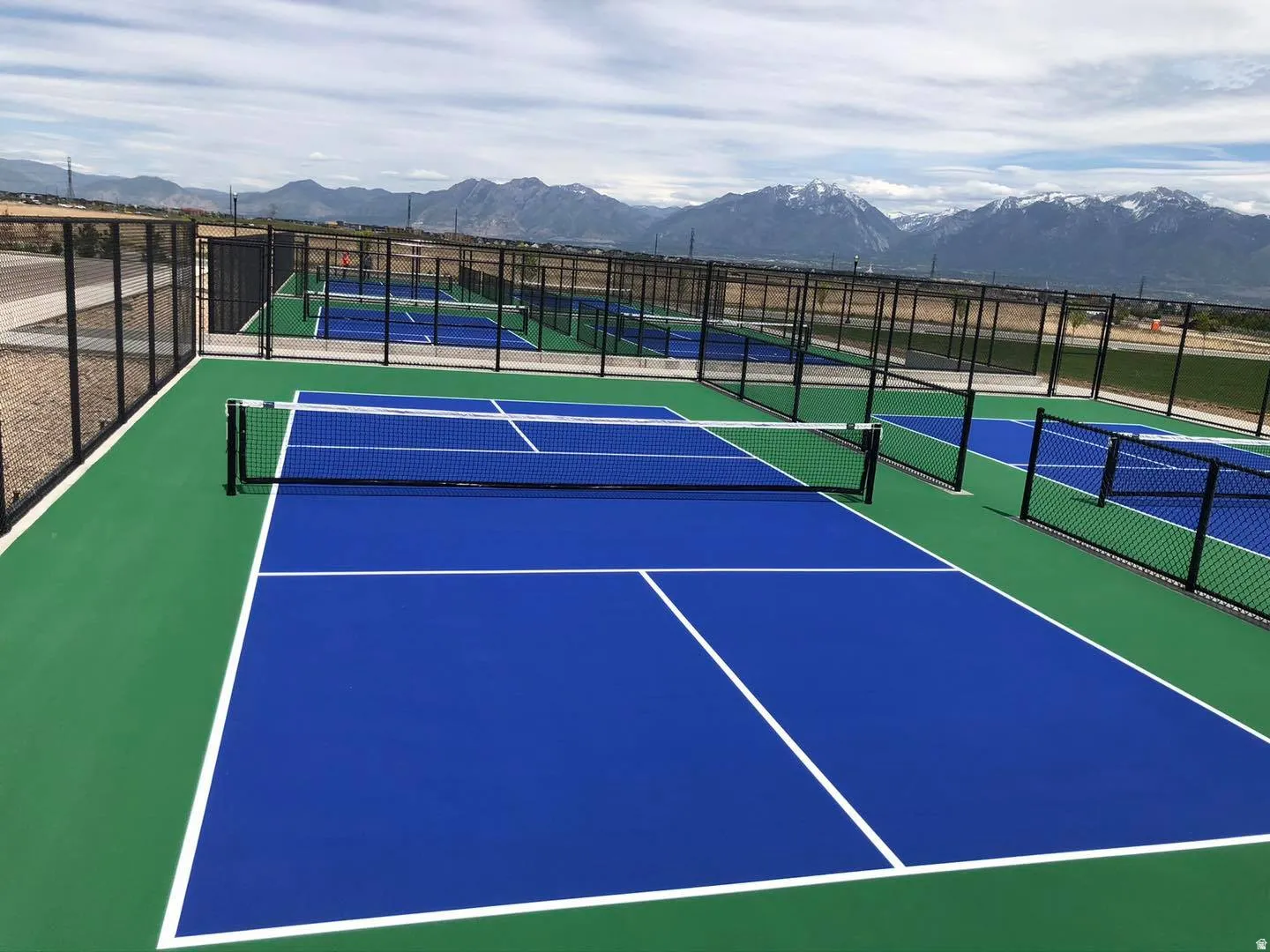 Multiple pickleball courts with blue and green surfaces, black chain-link fencing, and a mountain backdrop