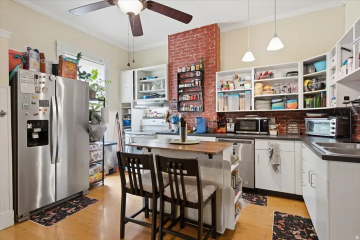 Exposed brick accents, light wood-finish flooring, white cabinetry, stainless steel appliances, and a ceiling fan with integrated lighting