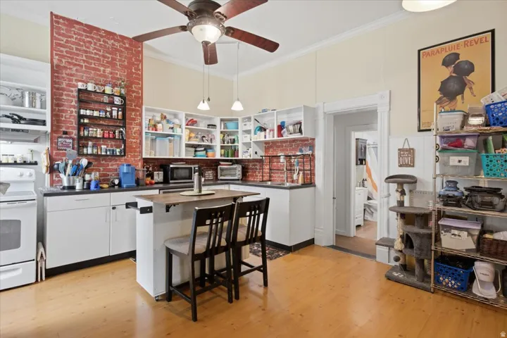 Hardwood-style flooring, exposed brick accent wall, white cabinetry with dark countertops, built-in shelving, and a ceiling fan with integrated lighting