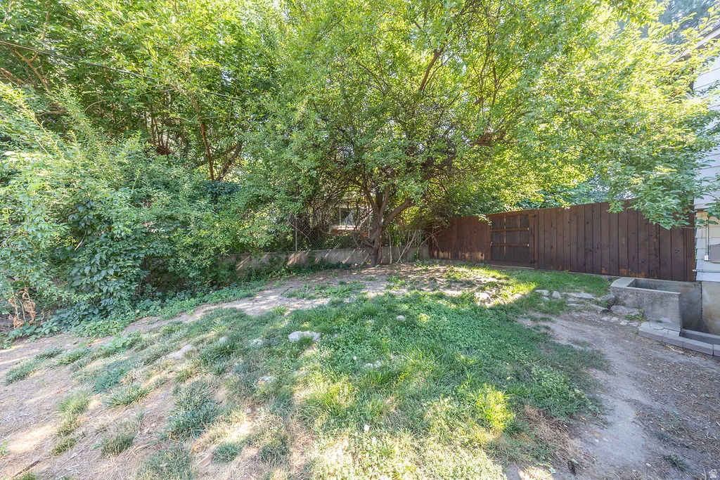 Sloping backyard featuring mature trees, partial grass cover, a full-height wood fence, and a concrete foundation element