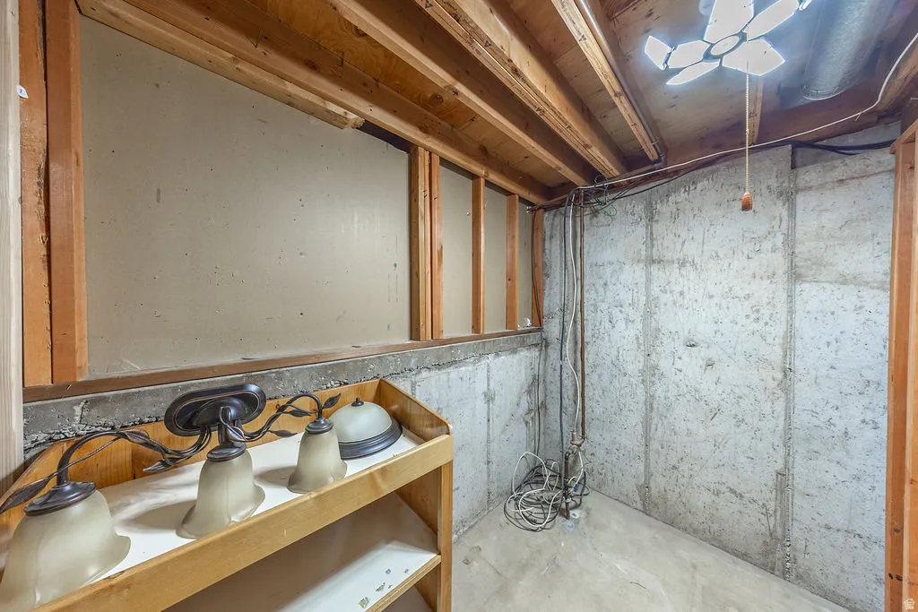 Unfinished basement area featuring exposed wooden ceiling joists, concrete walls, a small recessed window opening, and a built-in wooden shelf