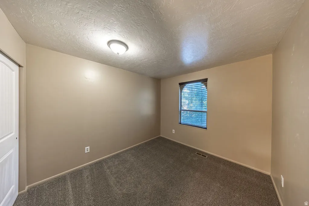Interior room featuring neutral wall paint, textured ceiling, a single window with blinds, and dark gray carpet flooring