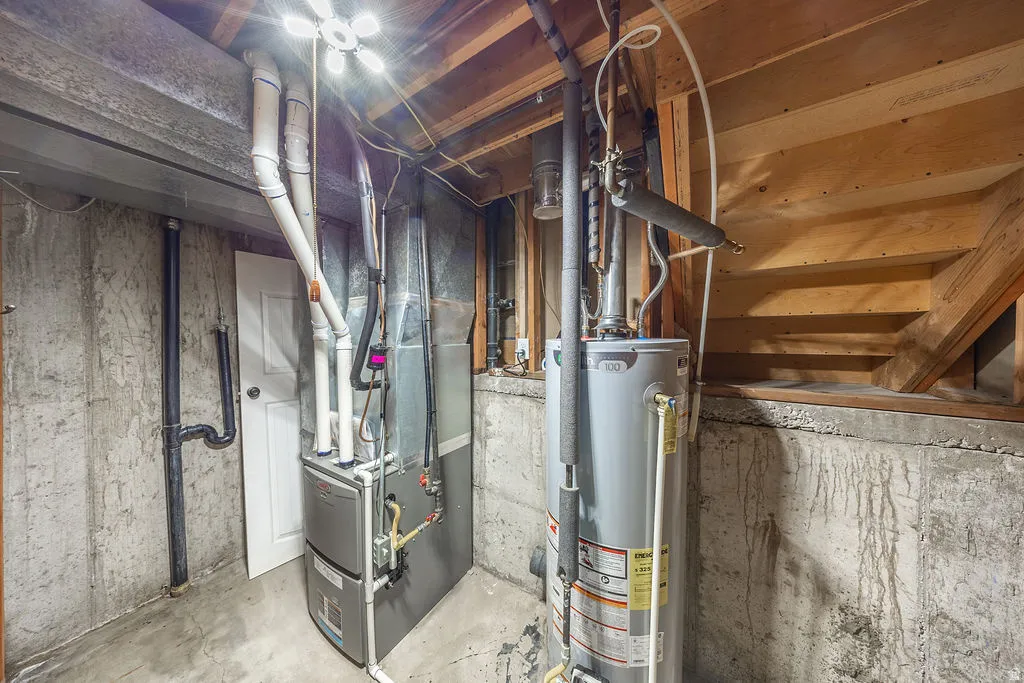 Utility area featuring a water heater and furnace with exposed concrete walls, wood ceiling joists, and a finished white door