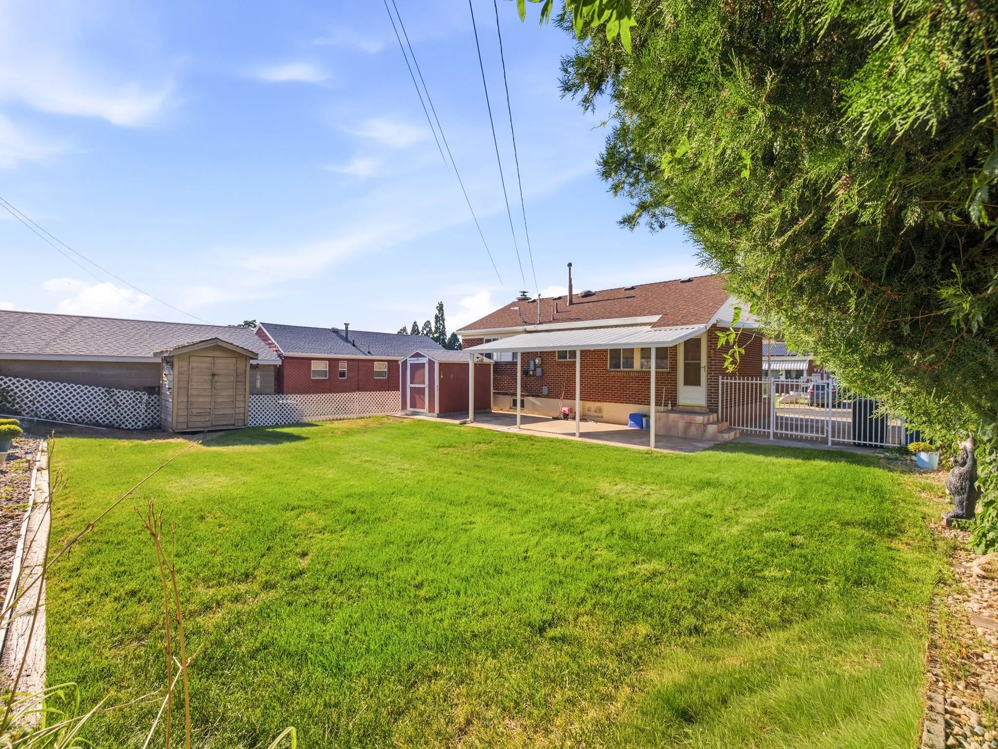 Expansive grassy yard with a covered patio, brick exterior, two storage sheds, and white metal fencing