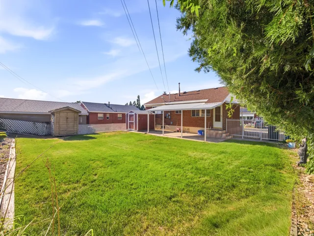 Expansive grassy yard with a covered patio, brick exterior, two storage sheds, and white metal fencing
