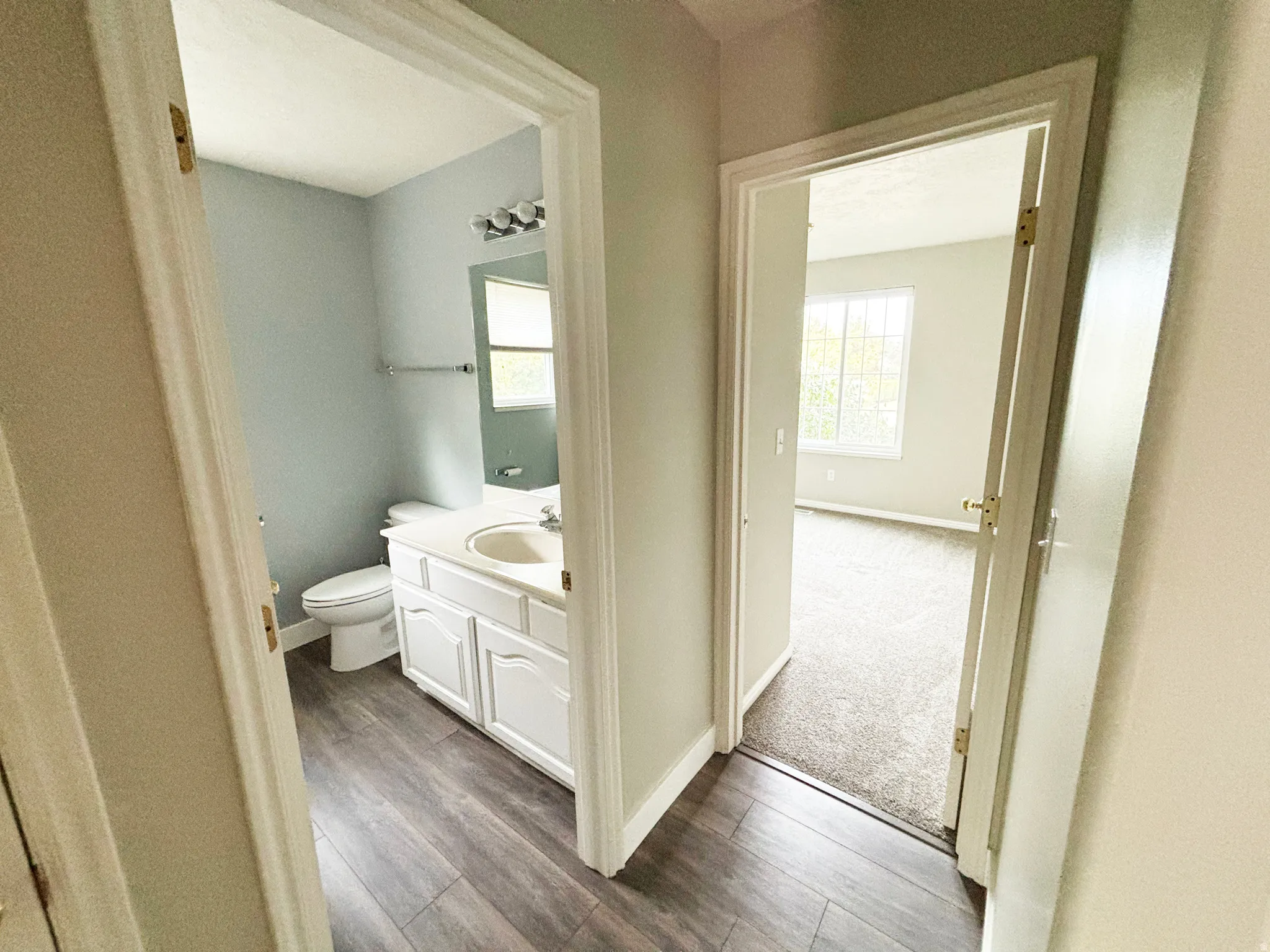 Hallway with wood-finish flooring leading to a full bathroom featuring white vanity cabinetry, dual sink setup, and tile flooring, with a secondary bedroom or living space visible through the adjacent doorway