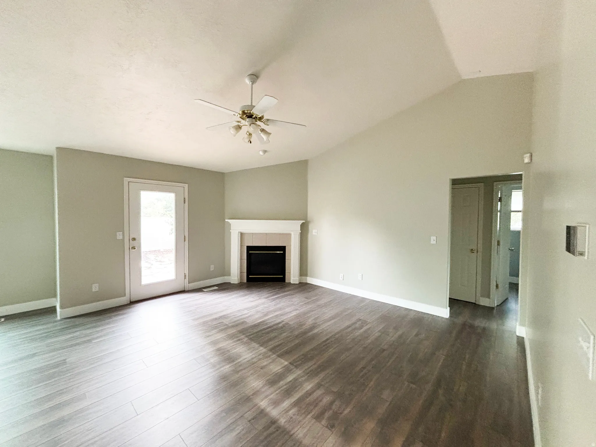Spacious living area featuring wood-finish flooring, a fireplace with white surround and mantel, vaulted ceiling with ceiling fan, and multiple windows providing natural light
