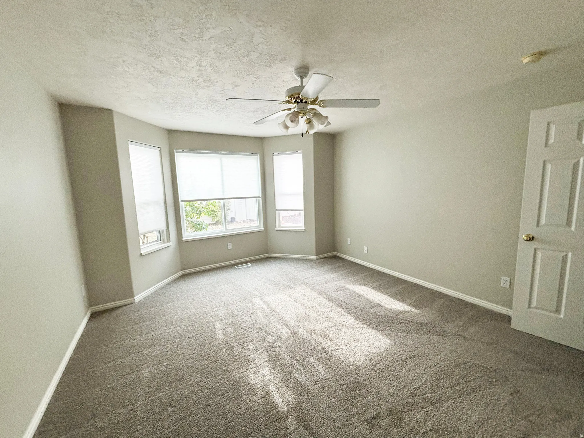 Bay window with multiple windows, textured carpeting, neutral wall color, and a ceiling fan with integrated lighting