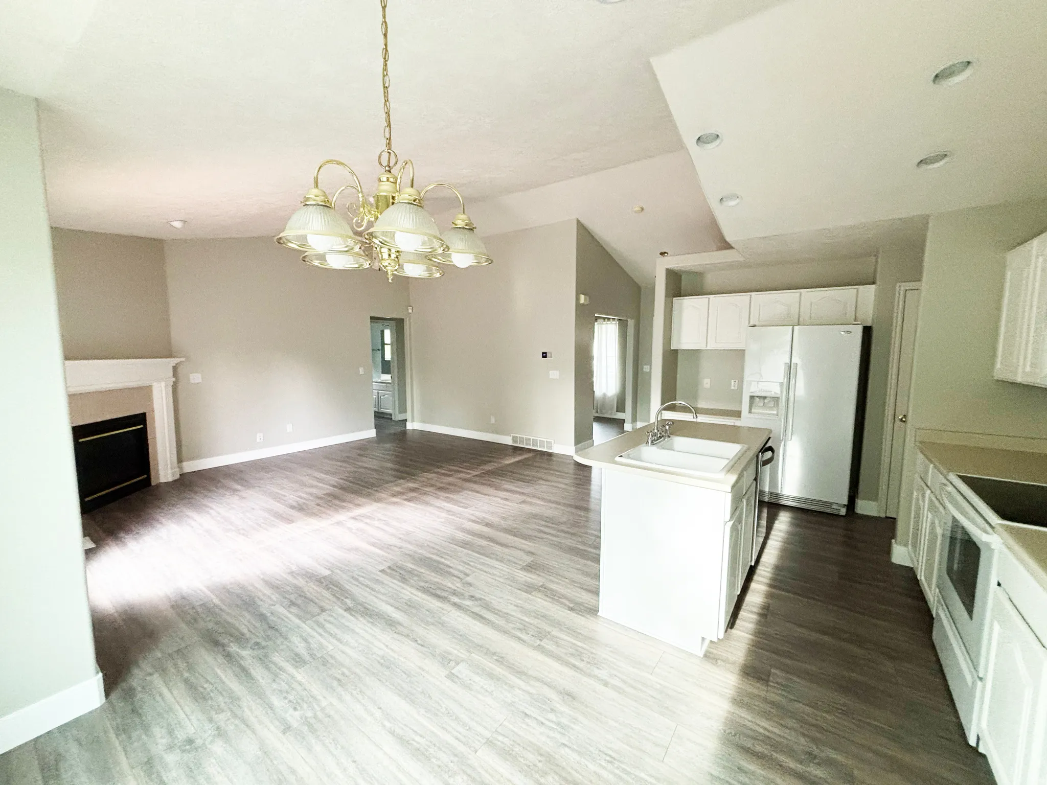 Open-concept living and kitchen area featuring wood-finish flooring throughout, a brass chandelier, white cabinetry with stainless steel appliances, an island with seating, and recessed ceiling lighting