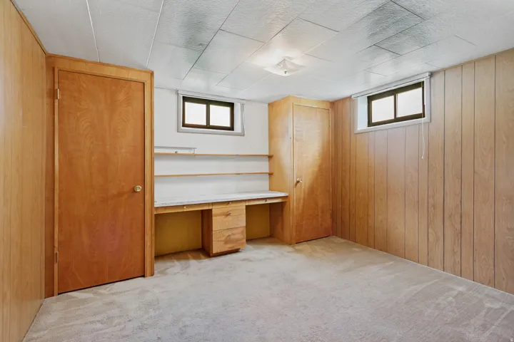 Built-in desk with shelving, wood-paneled walls, two windows, and textured ceiling tiles