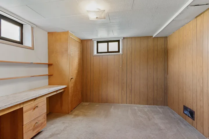 Built-in desk with shelving, wood-paneled walls, carpeted flooring, and a flush mount ceiling light