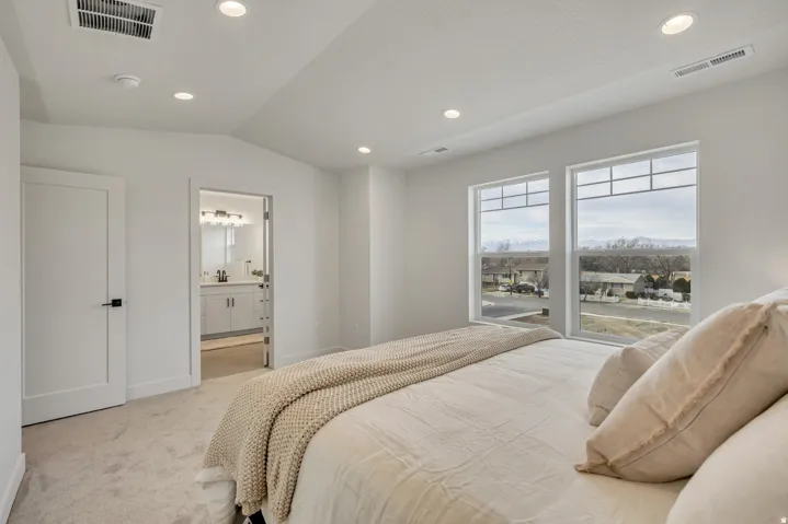 Bright primary bedroom showcasing floor-to-ceiling windows with expansive natural light, neutral wall finishes, recessed ceiling lighting, hardwood-finish flooring, an ensuite bathroom with dual vanity and chrome fixtures, and a walk-in closet with built-