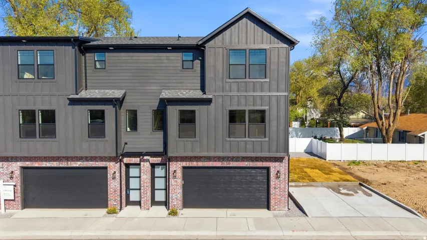 Modern duplex featuring dual two-car garage bays with dark panel doors, brick facade foundation, and contemporary gray fiber cement siding