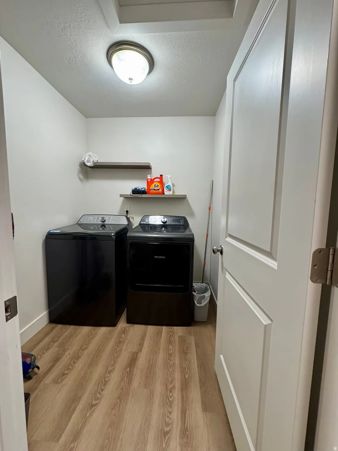 Dedicated laundry area featuring wood-finish flooring, two floating shelves, and a ceiling-mounted light fixture