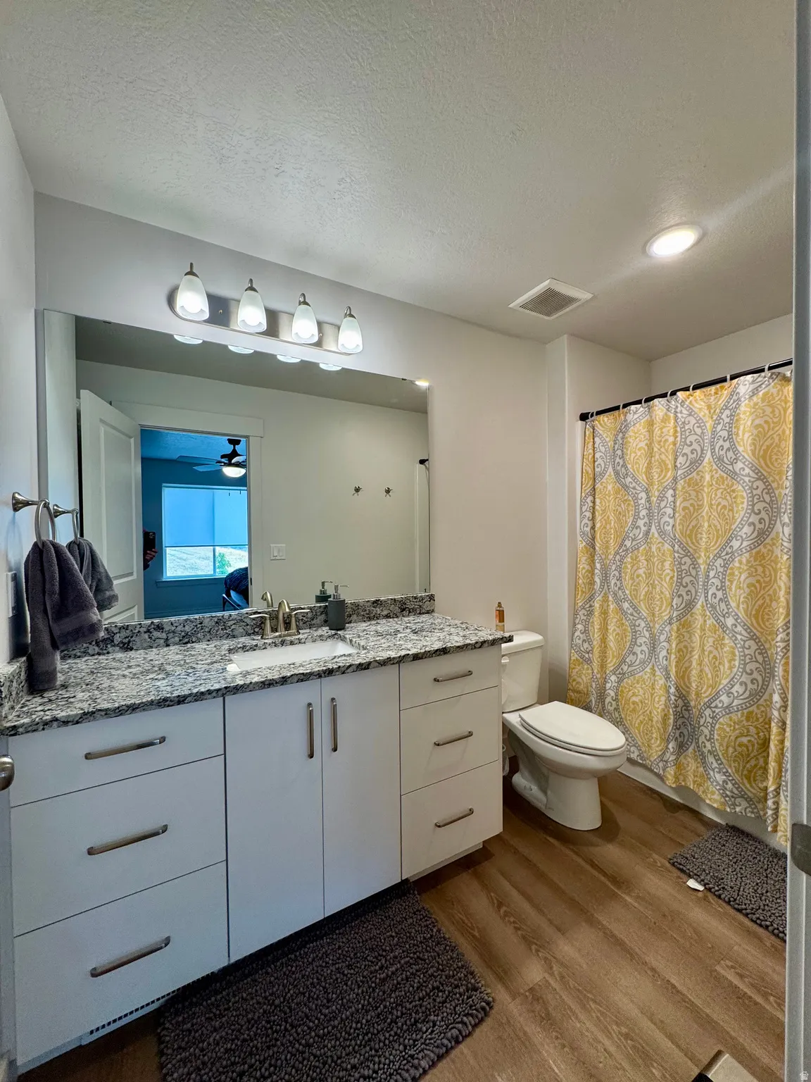 Vanity with granite-style countertop, white cabinetry with brushed nickel hardware, expansive wall mirror, and wood-finish flooring