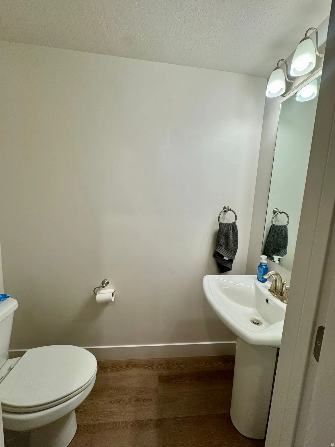 Pedestal sink with a brushed nickel faucet, wall-mounted vanity light fixture, wood-finish flooring, and a white toilet