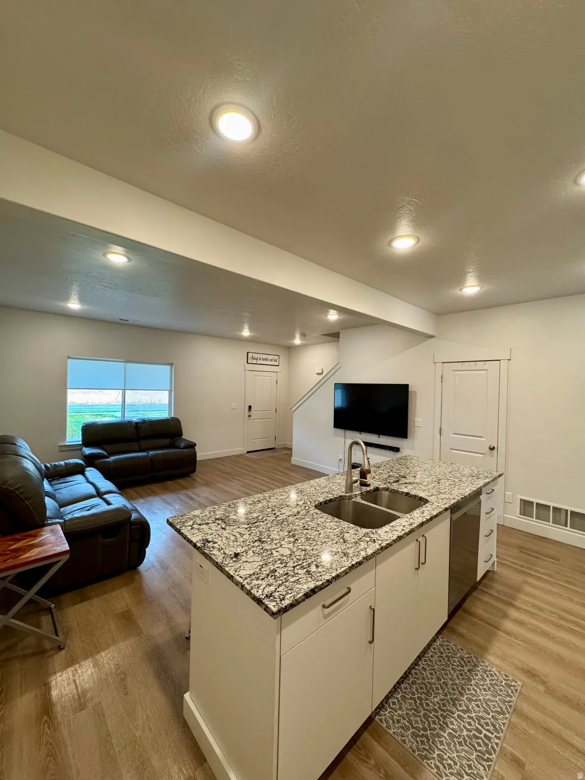 Open-concept living space featuring wood-finish flooring, recessed lighting, and a kitchen island with a double sink and granite-style countertop