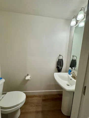 Pedestal sink with a brushed nickel faucet, wall-mounted vanity light fixture, wood-finish flooring, and a white toilet