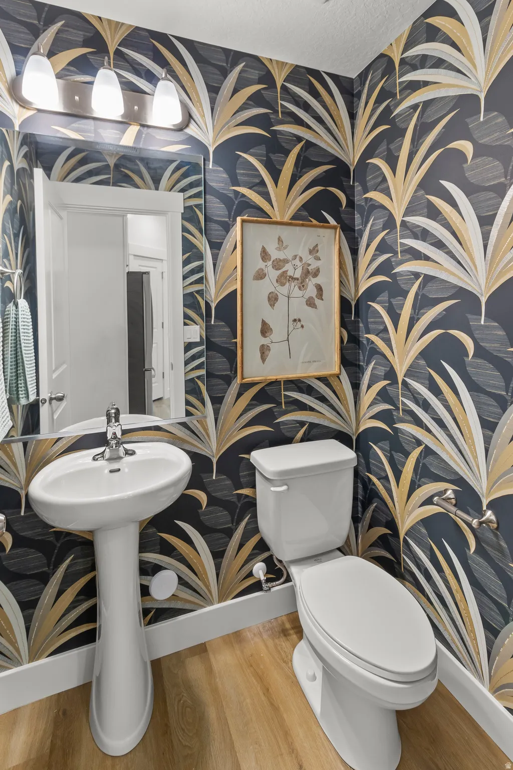 Pedestal sink with chrome faucet, toilet, wood-finish flooring, and wall-mounted vanity lighting