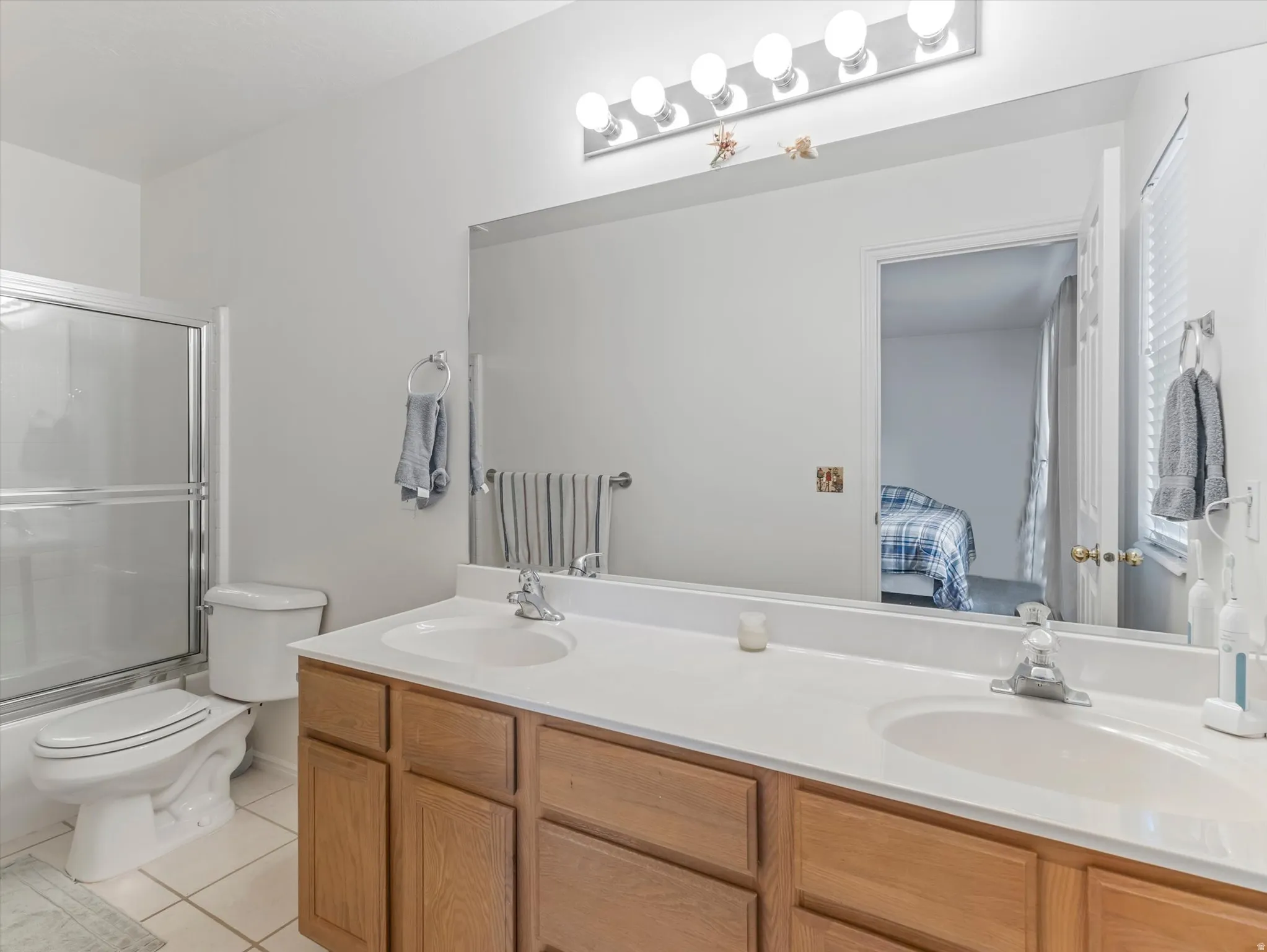 Dual vanity with white countertop and integrated sinks, wood-finish cabinetry, full-width mirror, and a shower-over-tub combination with sliding glass doors