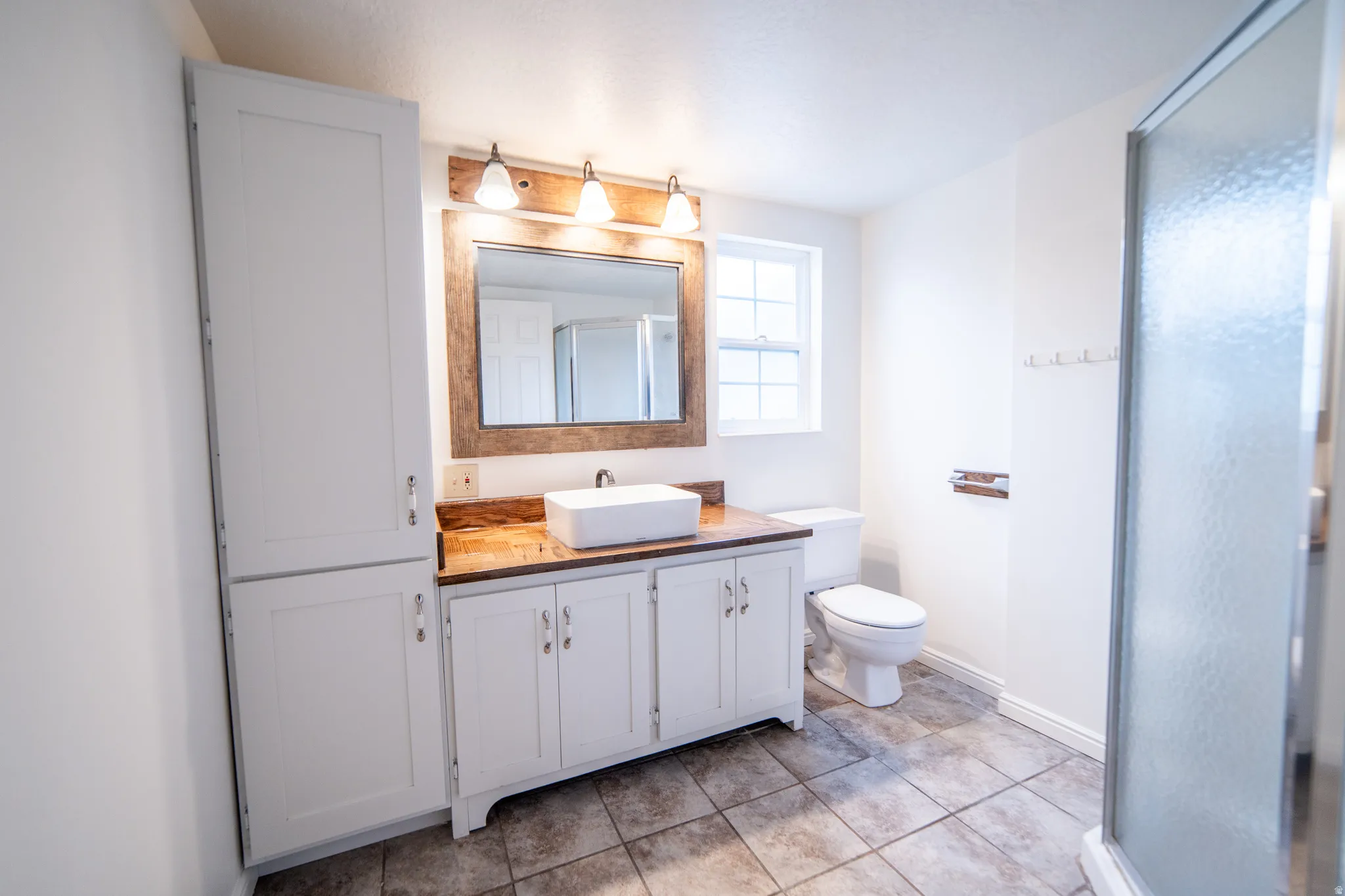 White shaker cabinetry, wood-finish countertop, vessel sink, tiled flooring, and a glass-enclosed shower
