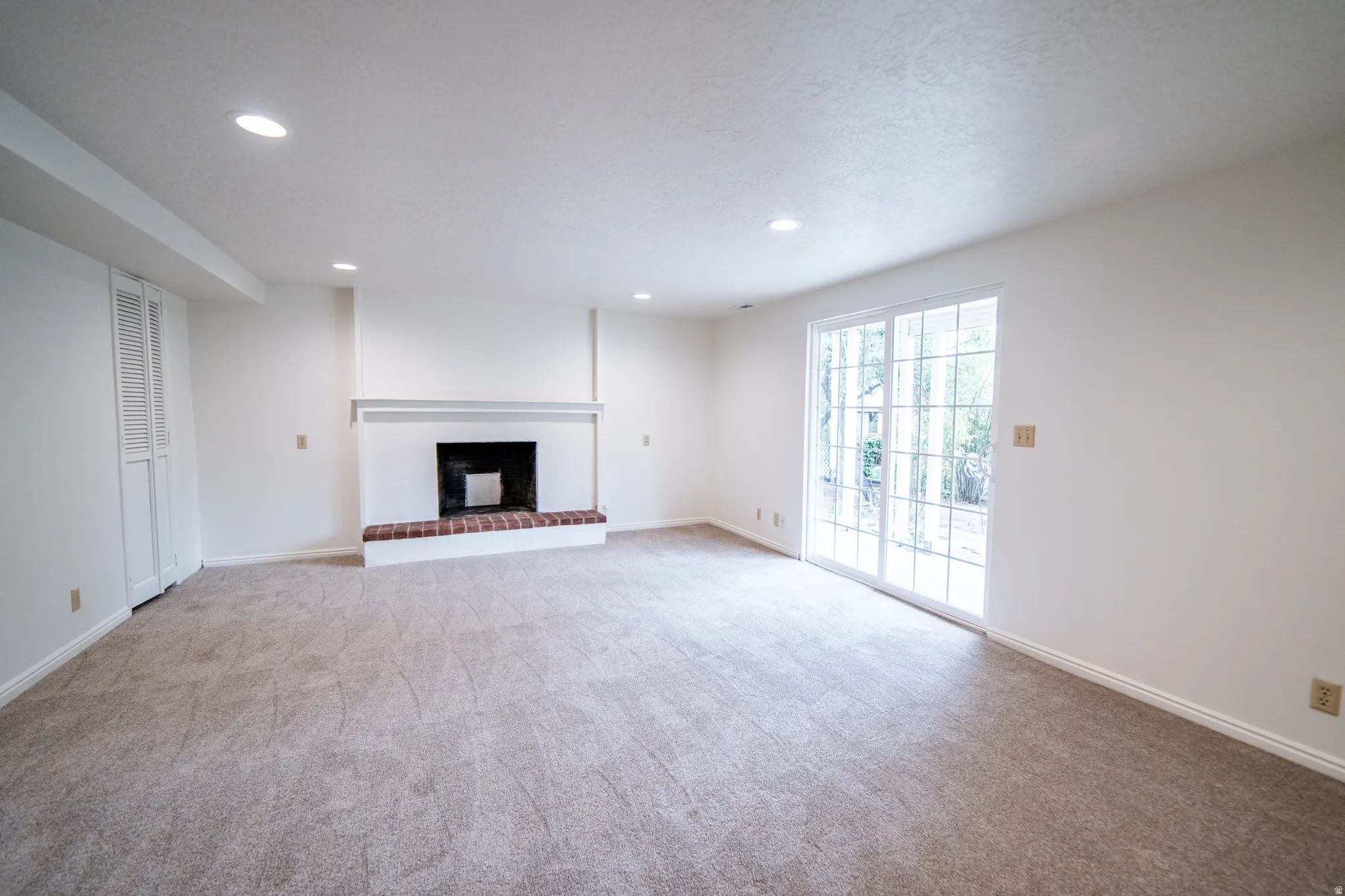 Recessed lighting illuminates a carpeted room featuring a fireplace with brick hearth and white mantel, and sliding glass doors leading to the backyard.