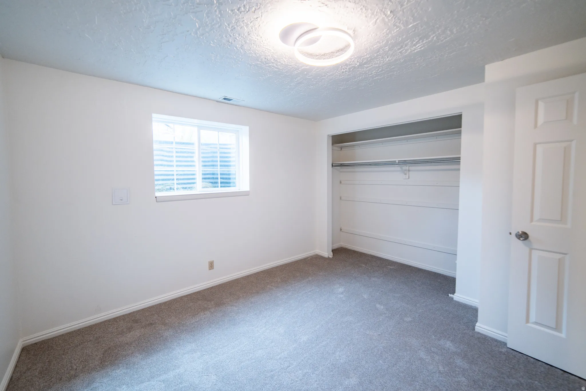 Neutral wall finishes with a single window, overhead flush-mount lighting, gray carpet, and a white closet with built-in shelving