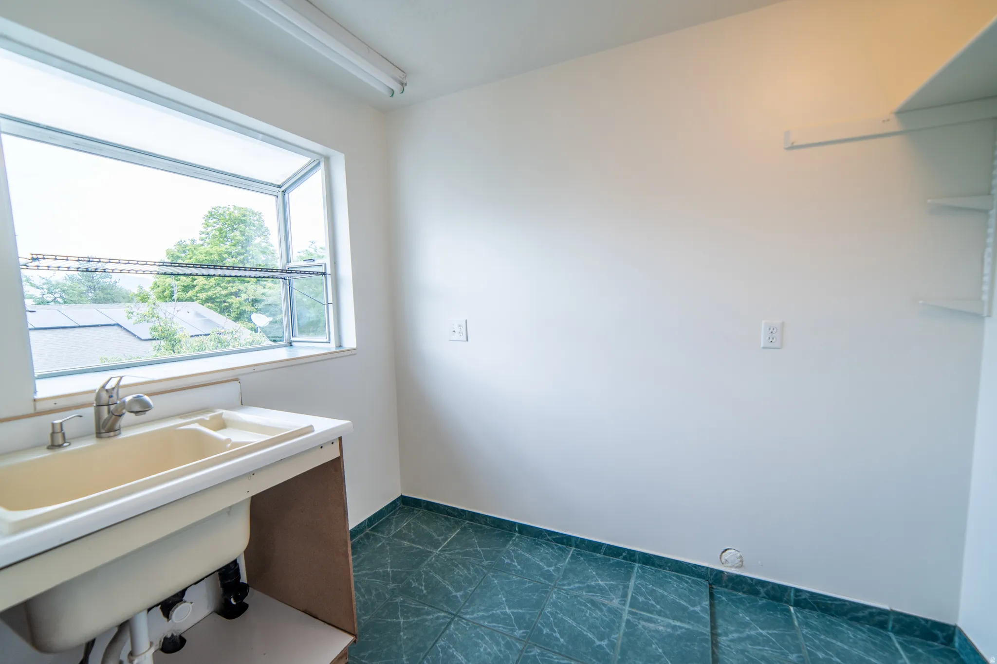 Utility sink with chrome faucet, large window with natural light, wall-mounted shelving, and patterned floor tile