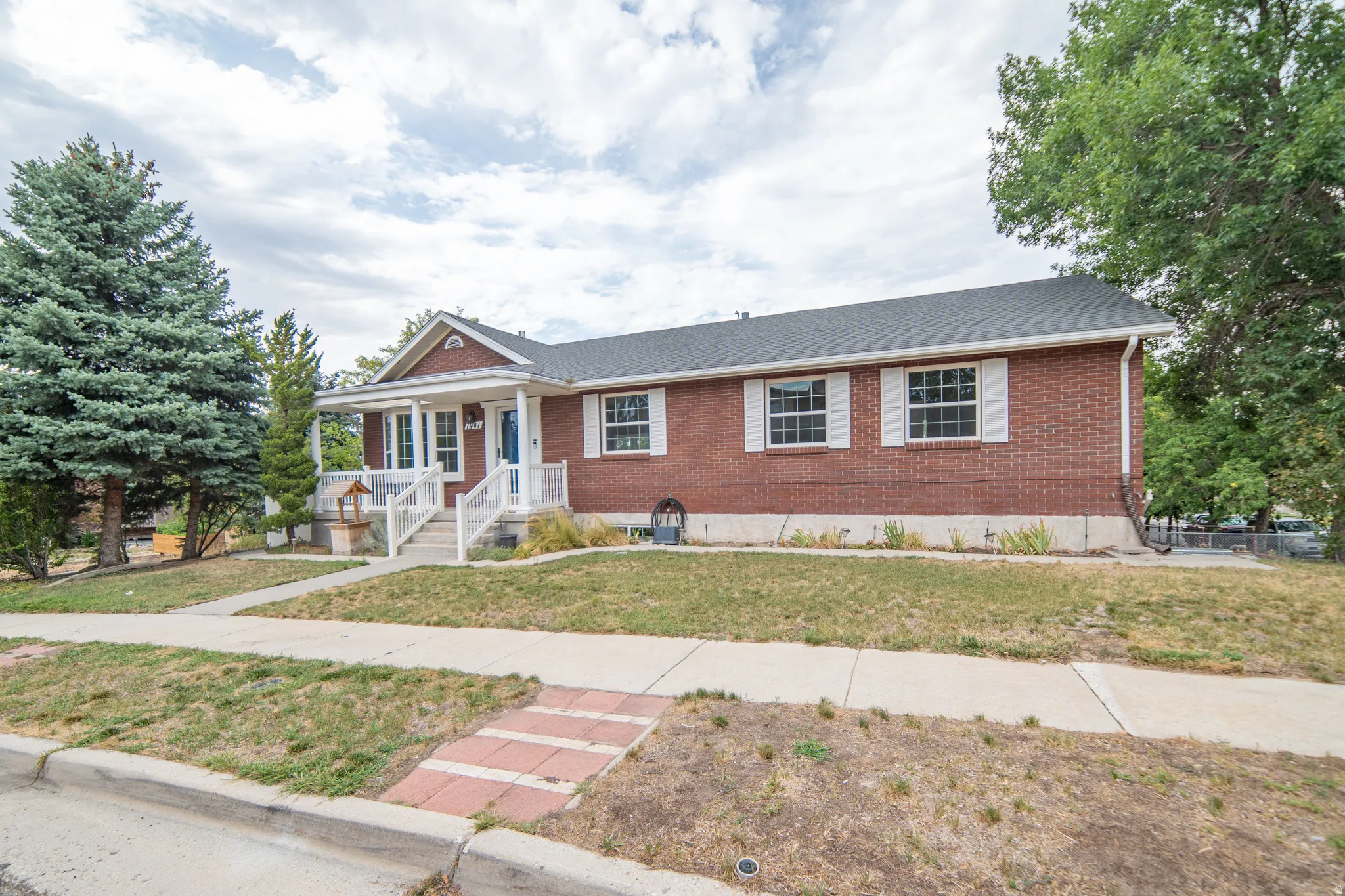 Brick exterior with a covered front porch featuring white railings, multiple mature trees, and a concrete walkway