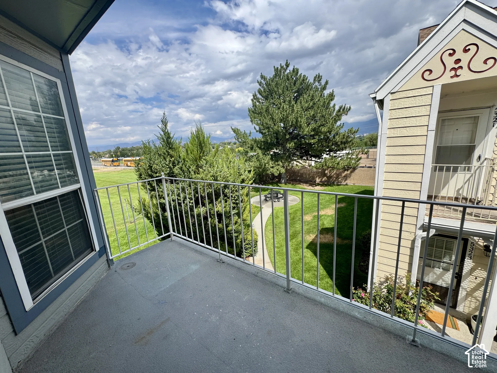 Private balcony with metal railings and a blue-gray floor, offering a green landscape with mature trees