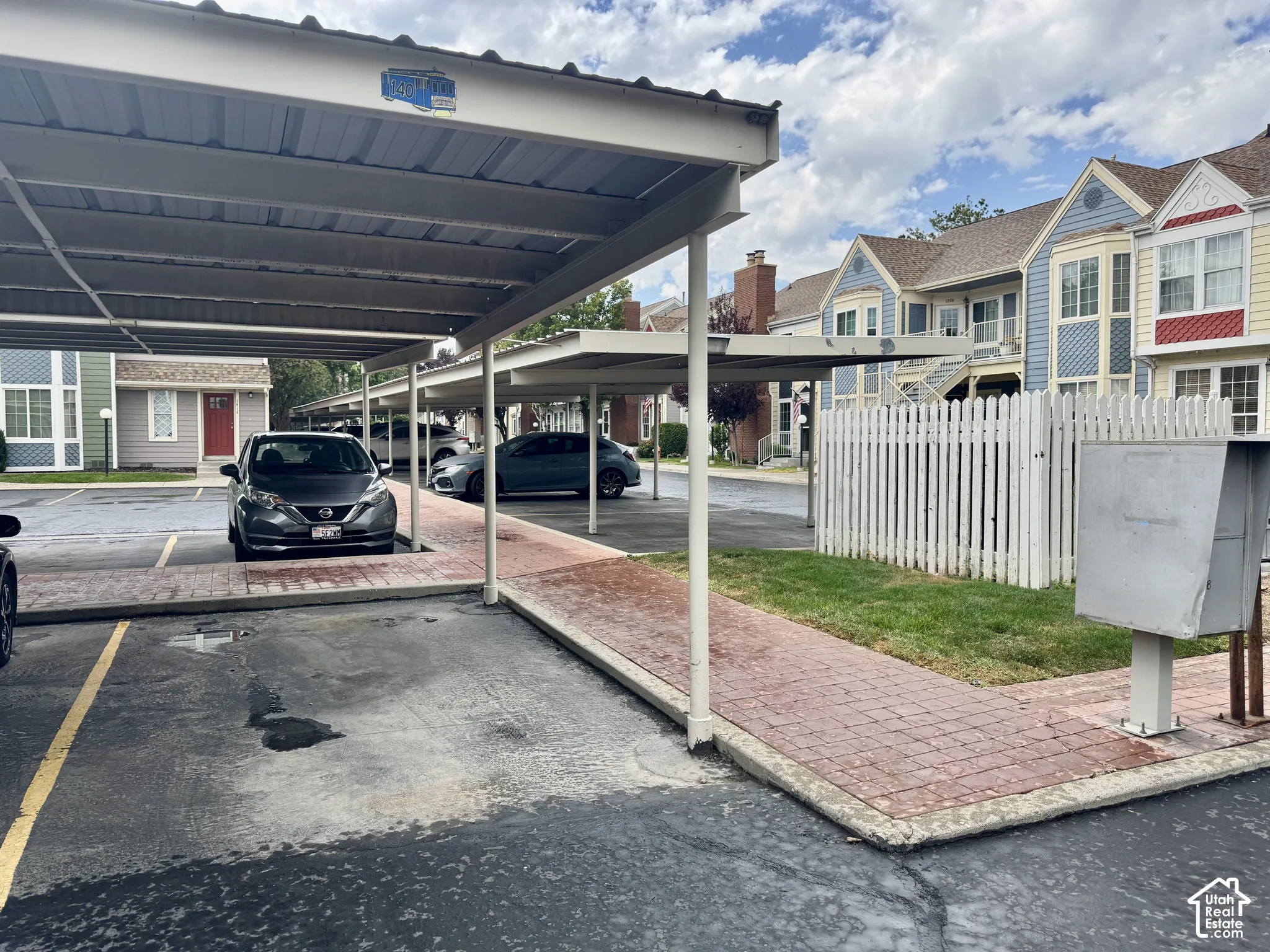 Covered parking structure with metal roofing, red brick-finish pathway, and painted wood picket fence