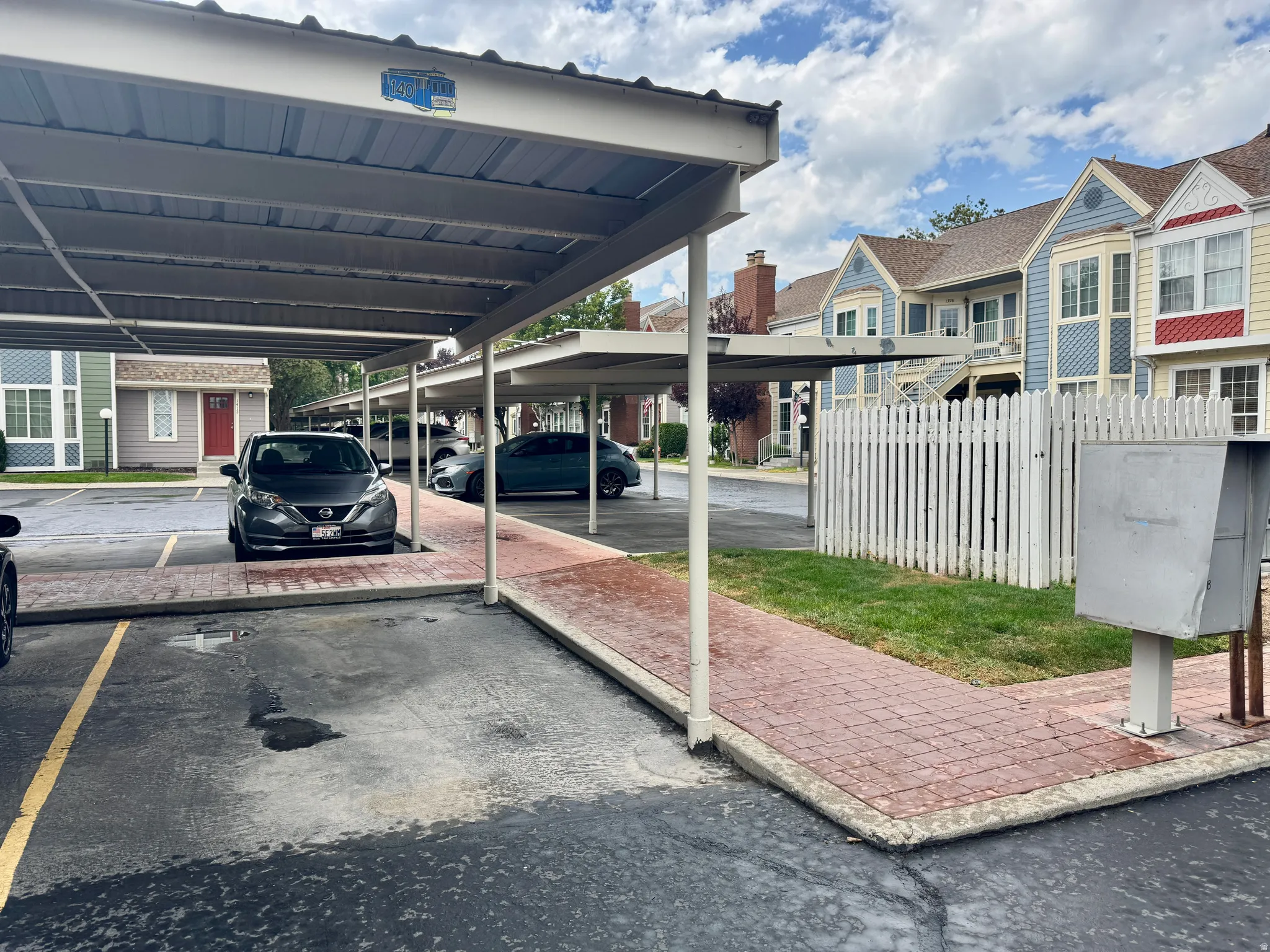 Covered parking structure with metal roofing, red brick-finish pathway, and painted wood picket fence