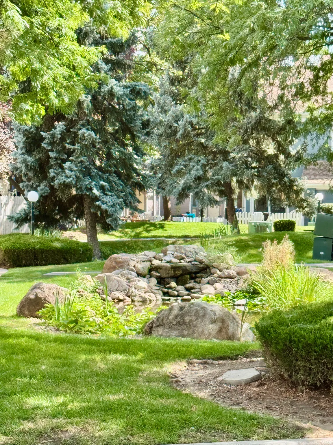 Landscaped grounds featuring a rock waterfall, pond with aquatic plants, diverse mature trees, and manicured shrubbery