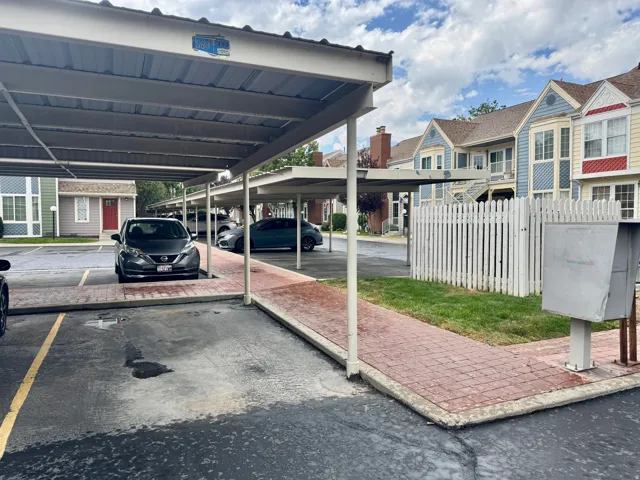 Covered parking structure with metal roofing, red brick-finish pathway, and painted wood picket fence