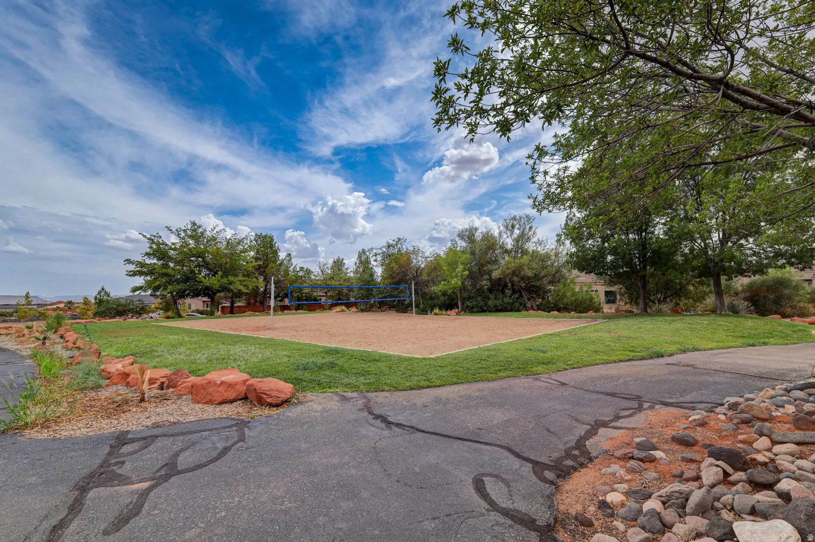 Sand volleyball court with net and boundary lines, surrounded by manicured lawn, established trees, and natural rock landscaping