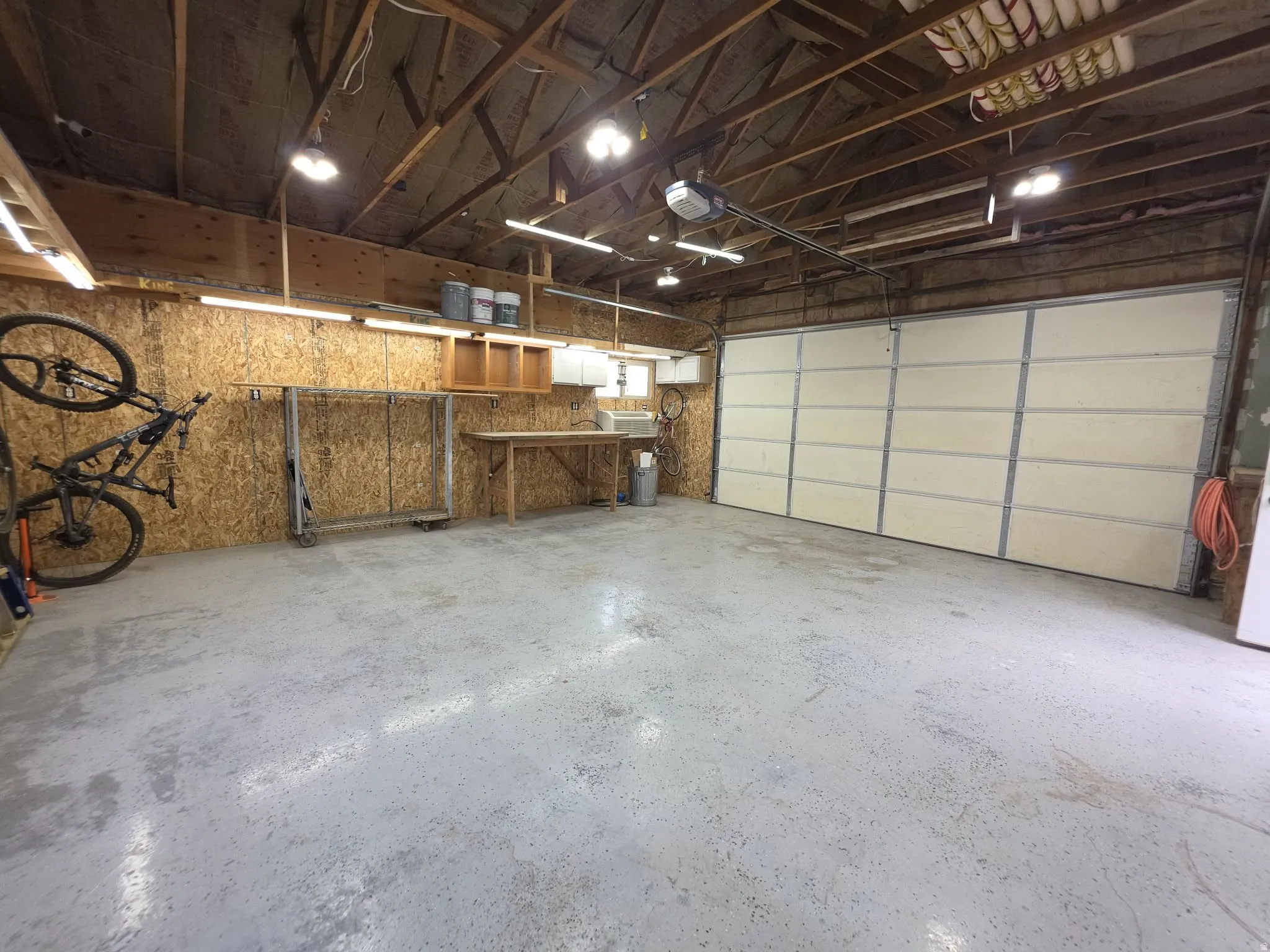 Spacious garage with an epoxy-coated concrete floor, exposed wood beam ceiling, multiple overhead light fixtures, a segmented garage door, and built-in workbench with cabinetry