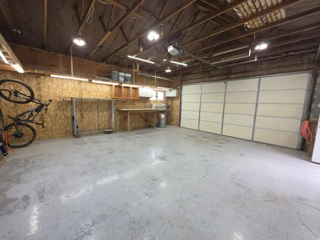 Spacious garage with an epoxy-coated concrete floor, exposed wood beam ceiling, multiple overhead light fixtures, a segmented garage door, and built-in workbench with cabinetry