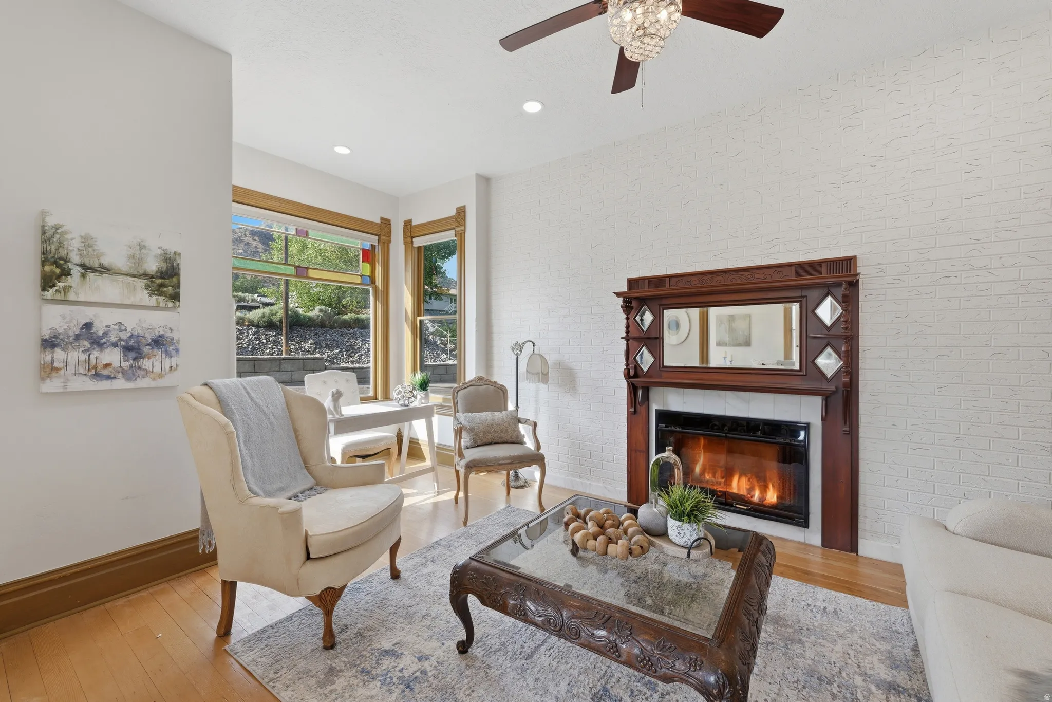 Hardwood flooring, white brick-finish wall, ornate wood fireplace surround with integrated mirror, crystal chandelier with ceiling fan, and large windows with stained glass accents