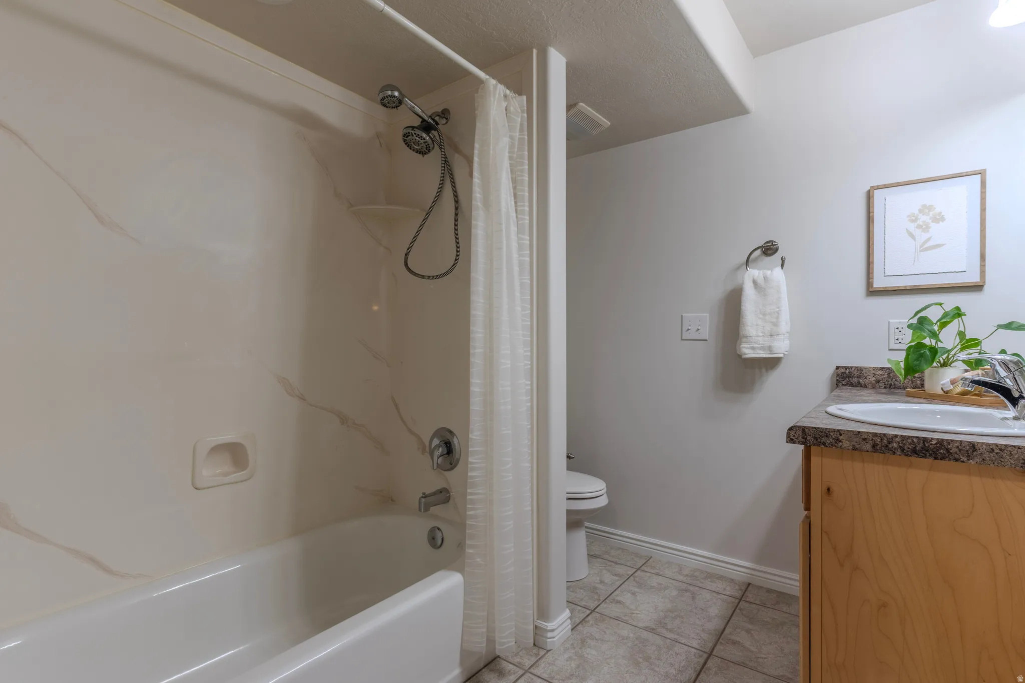 Tub and shower combination with a textured surround, light wood-finish vanity with an integrated sink, and tiled flooring