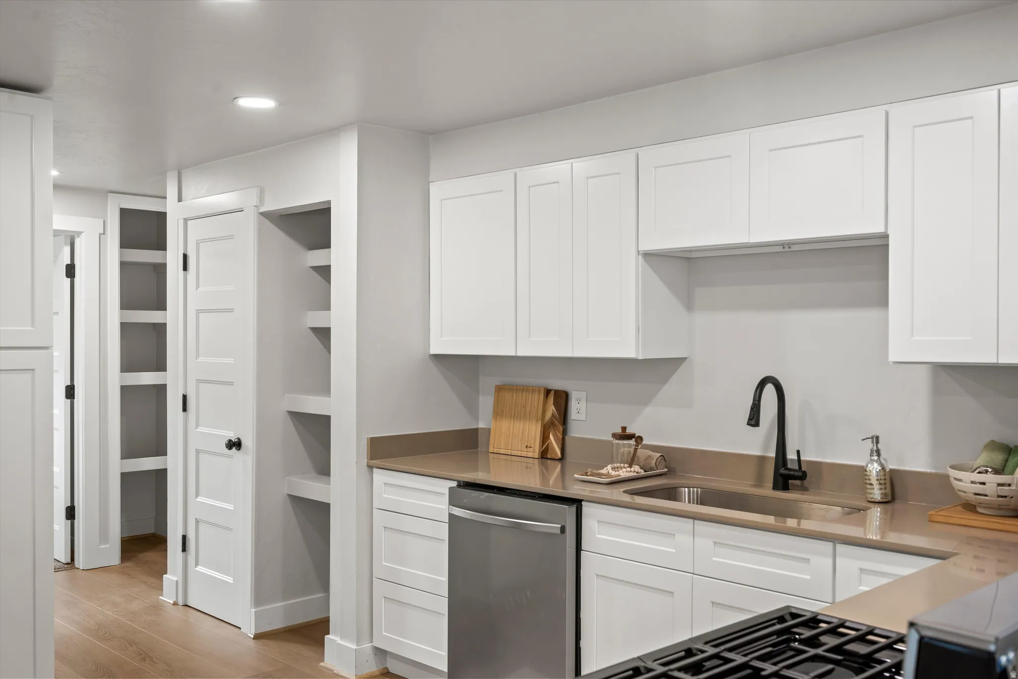 Kitchen featuring white shaker-style cabinetry, a stainless steel under-mount sink with a matte black faucet, light-toned countertops, and a built-in dishwasher