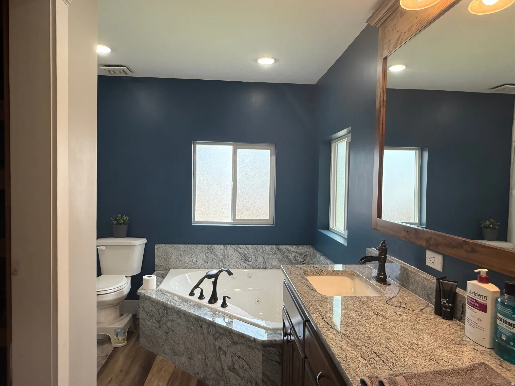 Built-in jetted tub with stone-finish surround, wood-finish flooring, recessed lighting, and a vanity with stone-finish countertop
