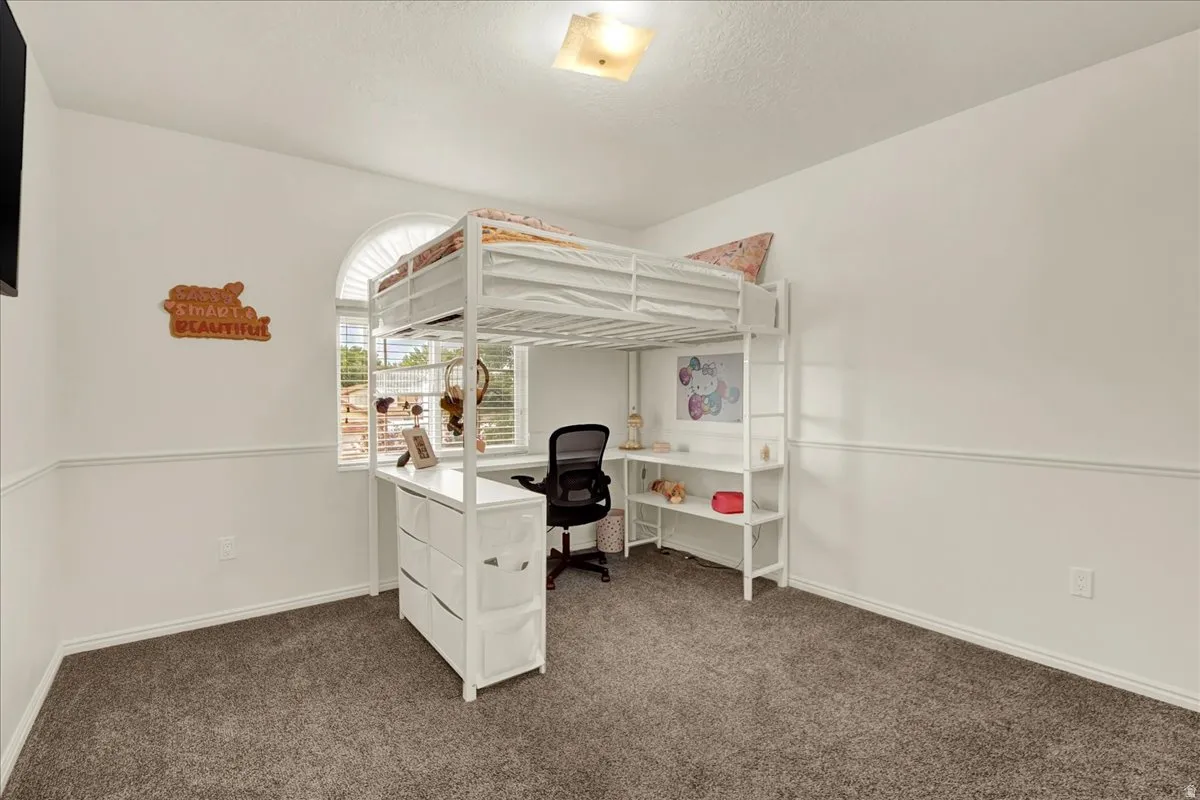 Versatile bedroom featuring a white metal loft bed with integrated desk and storage shelving, gray carpeting, recessed ceiling lighting, and ample wall space for personalization