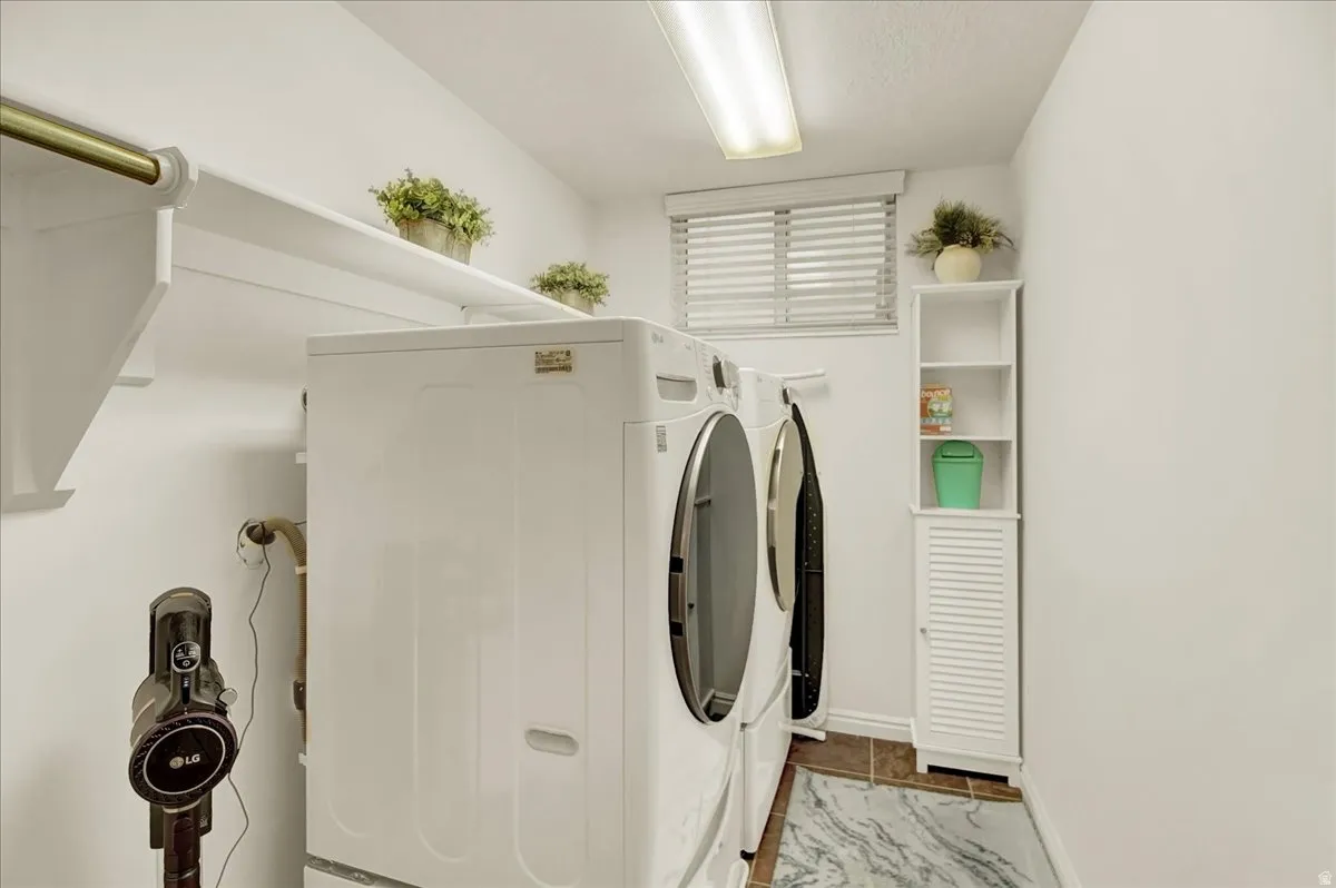 Bright, well-appointed laundry room featuring side-by-side front-load washer and dryer with stainless steel finishes, white cabinetry with louvered detailing, tile flooring, overhead shelving with decorative planters, and recessed lighting throughout