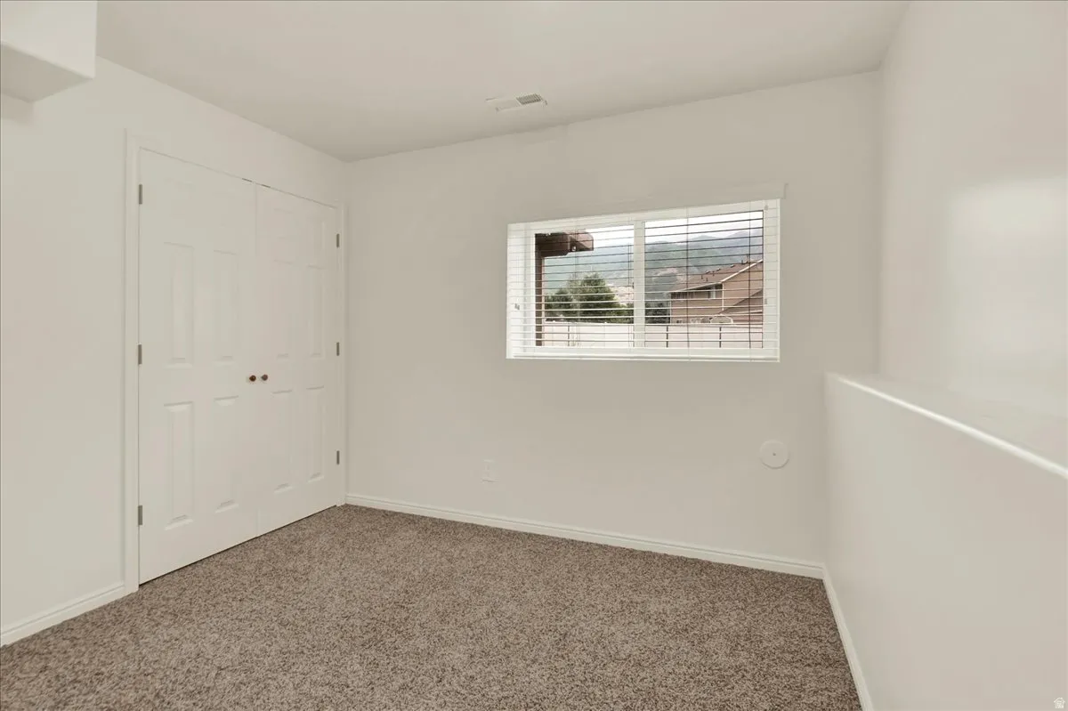 Bright secondary bedroom showcasing neutral wall finishes, carpet flooring, a double-door closet with white six-panel doors, and a horizontal window with exterior shutters allowing natural light