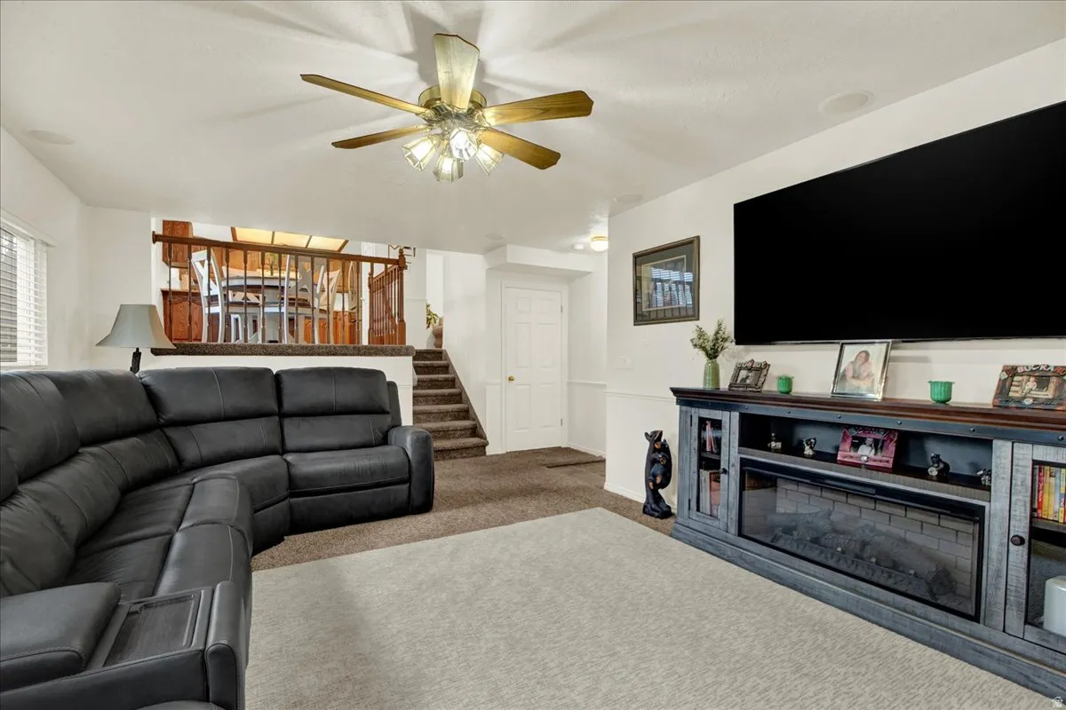 Spacious recreation room featuring a curved sectional sofa arrangement, wall-mounted flat-screen display, built-in media console with shelving and fireplace insert, ceiling fan with integrated lighting, neutral carpeting, and access to a wooden staircase