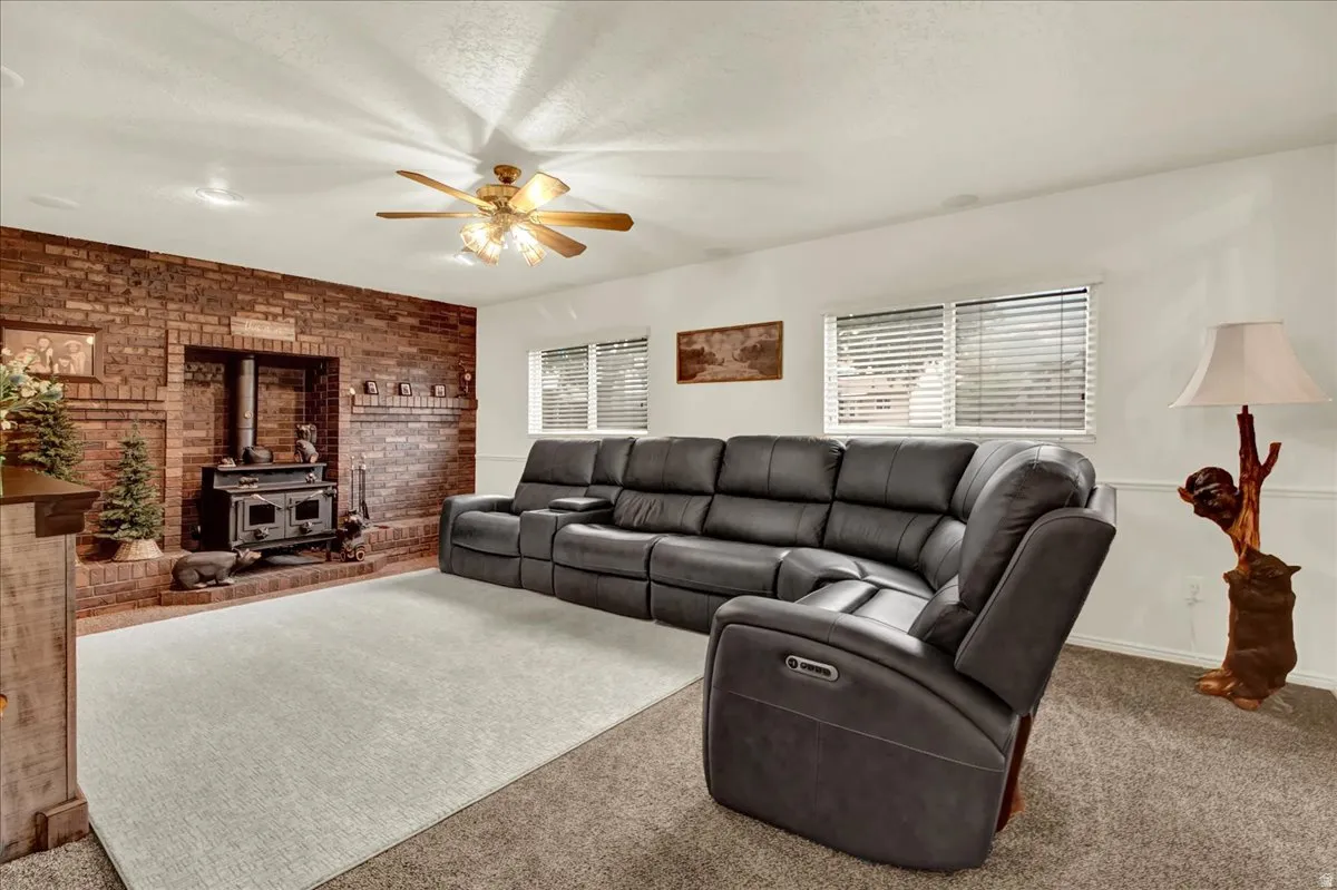 Spacious living room featuring a ceiling fan with light fixture, exposed brick fireplace with built-in hearth, expansive windows with plantation shutters, neutral carpeting, and recessed lighting throughout