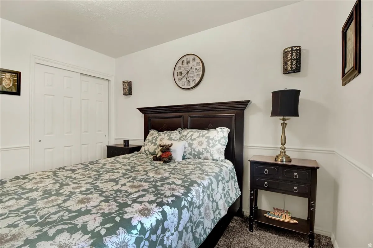 Bedroom featuring a dark wood headboard bed frame, coordinating two-drawer nightstand with brass-base table lamp, wall-mounted clock, framed wall décor, and carpeted flooring